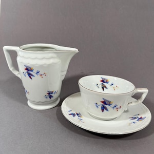 May include: A white porcelain teacup and saucer set with a matching creamer. The set features a gold trim and is decorated with blue, red, and yellow floral and geometric designs. The creamer has a scalloped rim.
