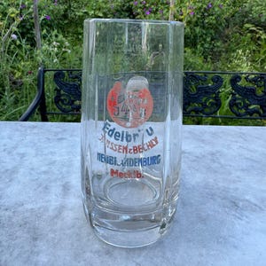 Könnte beinhalten: Ein klares, vintage Bierglas mit rechteckiger Form. Das Glas hat ein rundes Emblem mit roten und weißen Akzenten und den Text "Edelbr" und "Janssen & Bechly Neubrandenburg".
