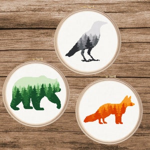 May include: Three embroidery hoops with cross-stitch designs of a bear, a fox, and a crow. Each animal is outlined in black and filled with a forest scene. The bear is green, the fox is orange, and the crow is grey.