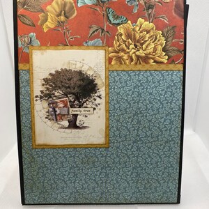 Op de afbeelding: Een scrapbook met een zwarte omslag en een bloemenpatroon. De voorkant heeft een boom met de woorden "family tree" op een bord dat aan een tak hangt. De achtergrond is een rood en blauw bloemenpatroon.