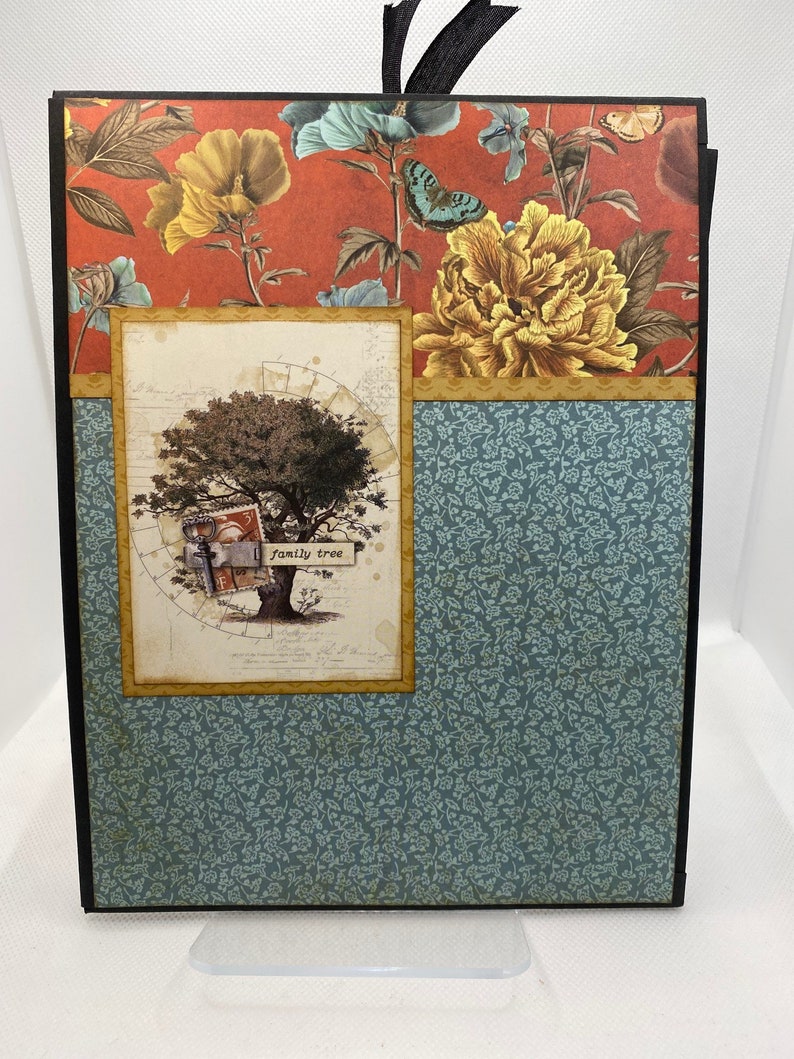 Op de afbeelding: Een scrapbook met een zwarte omslag en een bloemenpatroon. De voorkant heeft een boom met de woorden "family tree" op een bord dat aan een tak hangt. De achtergrond is een rood en blauw bloemenpatroon.