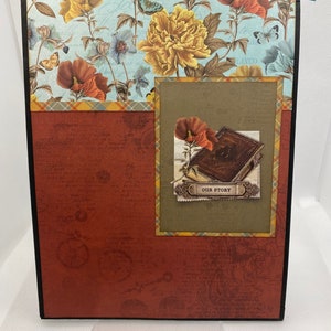 Op de afbeelding: Een rood scrapbook met een zwarte rand en een bloemenpatroon aan de voorkant. Het scrapbook heeft een kleiner venster met een bruine achtergrond en een bloemenontwerp. Het venster heeft een boek met de tekst "Our Story" erop.
