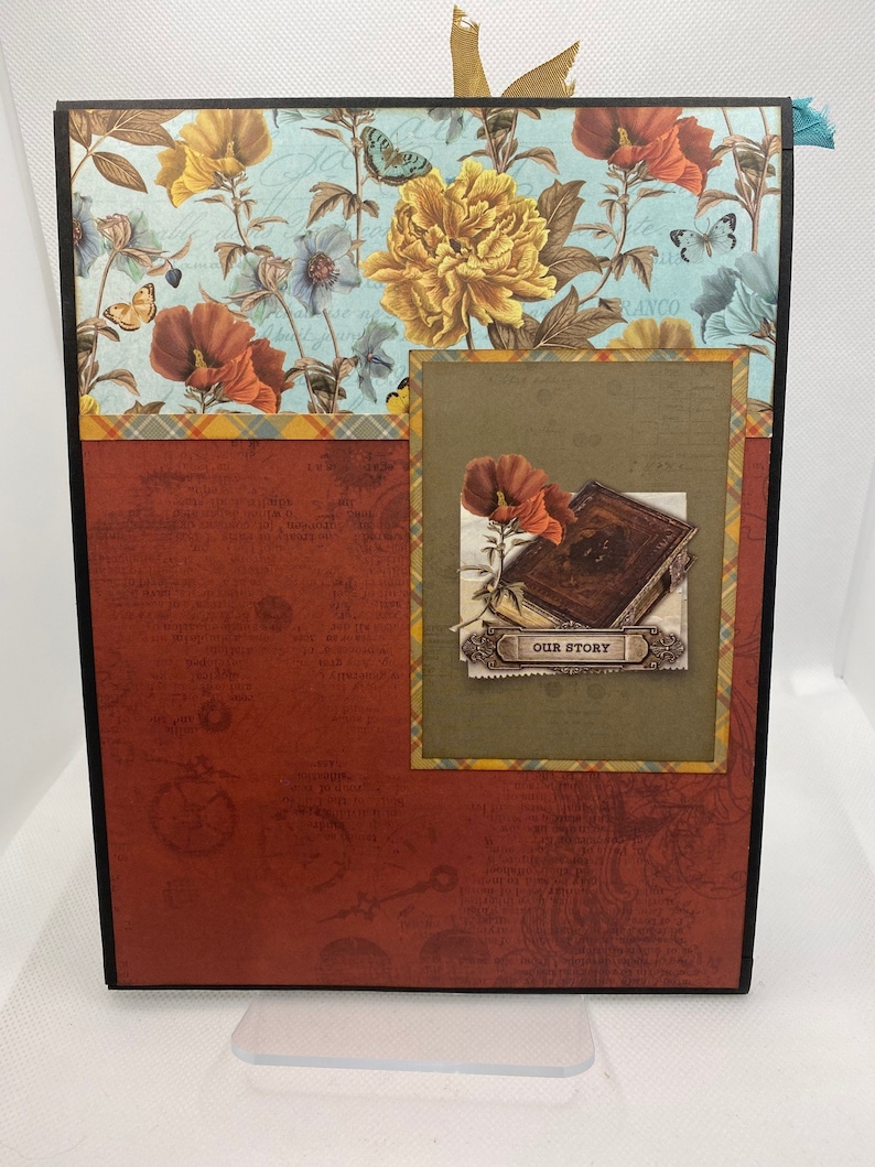 Op de afbeelding: Een rood scrapbook met een zwarte rand en een bloemenpatroon aan de voorkant. Het scrapbook heeft een kleiner venster met een bruine achtergrond en een bloemenontwerp. Het venster heeft een boek met de tekst "Our Story" erop.