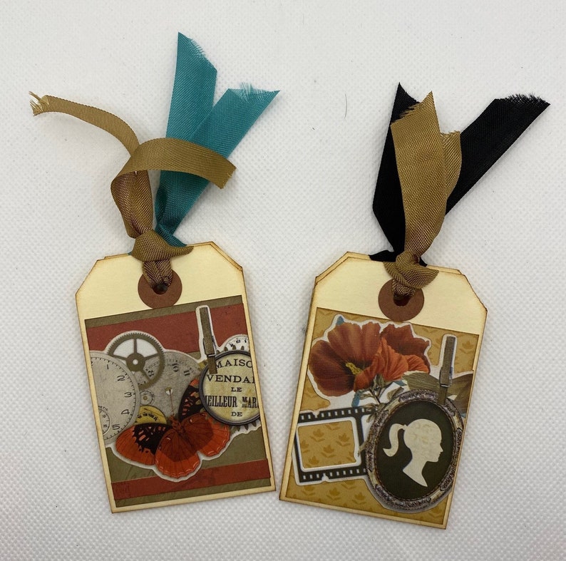 Op de afbeelding: Twee decoratieve labels met een bruin lint en een bruine papieren achtergrond. Een label heeft een vlinder, tandwielen en de tekst "Maison Vendale Le Meilleur March&eacute; de". Het andere label heeft een rode bloem, een filmstrip en een silhouet van een vrouw.
