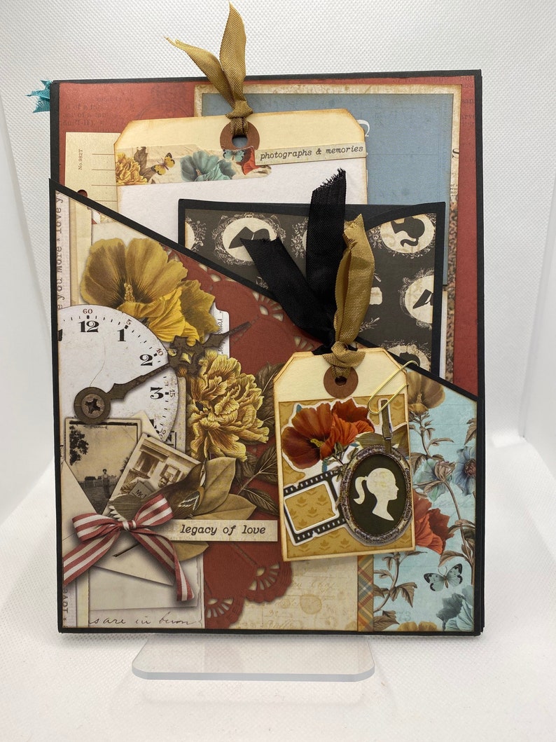 Op de afbeelding: Een scrapbook met een bruine, rode en zwarte omslag. De omslag heeft verschillende versieringen, waaronder bloemen, een klok, een label met de tekst "legacy of love" en een label met de tekst "photographs & memories".