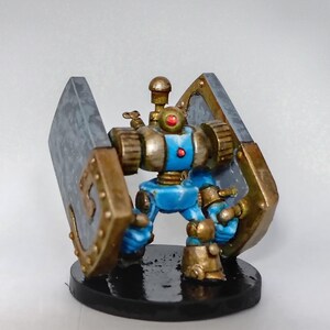 Può includere: Figurina di robot in miniatura con corpo blu, dettagli oro e argento e un occhio rosso. Il robot ha due grandi strutture a forma di scudo su entrambi i lati. Si trova su una base nera.