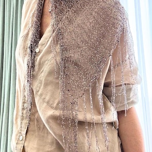 May include: A shimmering, light brown scarf with a delicate, net-like design and dangling fringe. The scarf is draped over a light brown button-down shirt, creating a layered effect. The scarf's texture and color suggest it is made of a lightweight material.