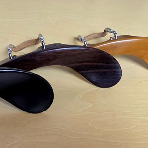 May include: Three violin shoulder rests with curved wooden bodies in black, dark brown, and light brown finishes. Each rest features adjustable metal hardware and a tan leather strap attached to the top, designed for ergonomic support.