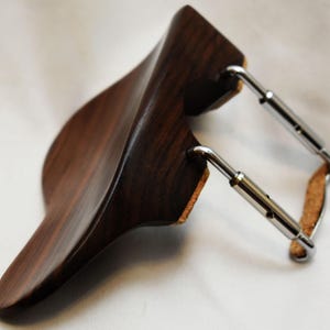 May include: A dark wood violin shoulder rest with a curved design. The rest has a polished finish and is connected to the instrument with adjustable metal hardware and cork padding.