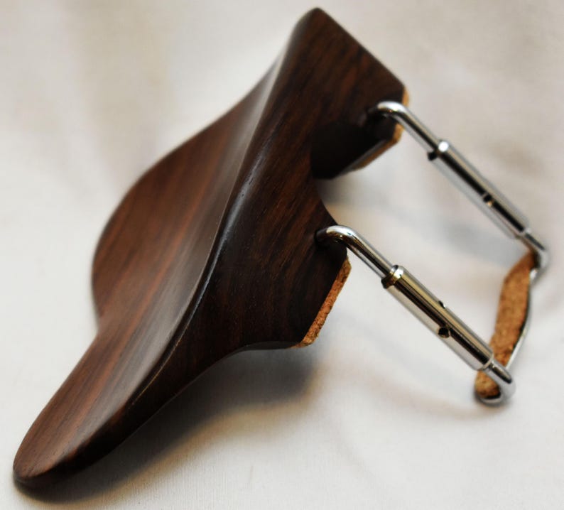 May include: A dark wood violin shoulder rest with a curved design. The rest has a polished finish and is connected to the instrument with adjustable metal hardware and cork padding.
