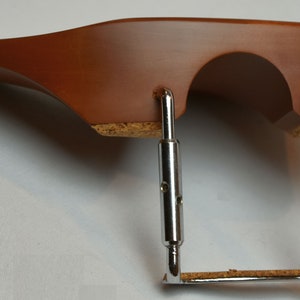 May include: A brown wooden violin shoulder rest with a curved design. It features a cork base and adjustable metal supports. The shoulder rest is designed to provide comfort and support for violin players.