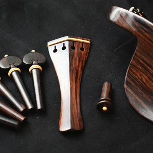 May include: A set of violin accessories made of dark wood with gold accents. The set includes a violin bridge, four tuning pegs, a tailpiece, and a chin rest.