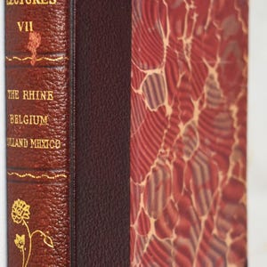 May include: A vintage book titled "Stoddard's Lectures VII" with a burgundy leather cover and gold floral detailing. The spine displays the title and the locations: The Rhine, Belgium, Holland, and Mexico. The book's cover has a marbled design.