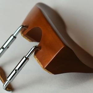 May include: A brown wooden violin shoulder rest with chrome metal brackets. The rest has a curved shape and is designed to fit comfortably on the shoulder. The brackets are adjustable for a secure fit. The rest is designed to elevate the violin for better playing posture.