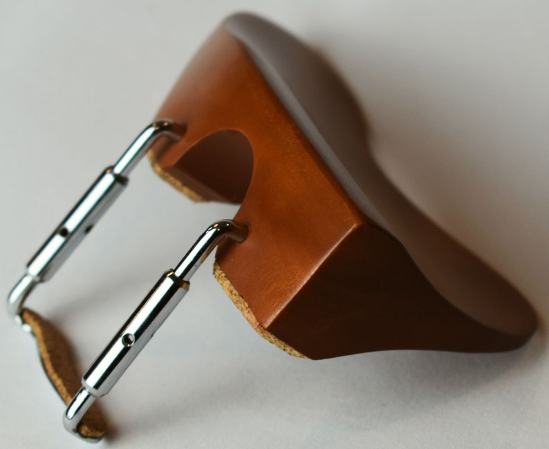 May include: A brown wooden violin shoulder rest with chrome metal brackets. The rest has a curved shape and is designed to fit comfortably on the shoulder. The brackets are adjustable for a secure fit. The rest is designed to elevate the violin for better playing posture.