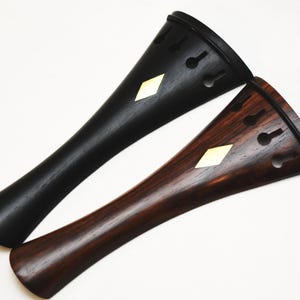 以下が含まれることがあります： 黒と濃い茶色のバイオリンのテールピース2つ。それぞれにひし形の象嵌が施されています。テールピースは先細りのデザインで、上部に鍵穴型の小さな切り欠きが並んでいます。茶色のテールピースには木目が見えます。