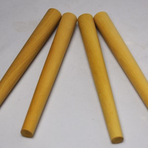 May include: Four wooden dowels, each with a light brown finish. The dowels are cylindrical, tapering slightly towards one end. They are arranged diagonally across a white surface, suggesting a craft or woodworking theme.