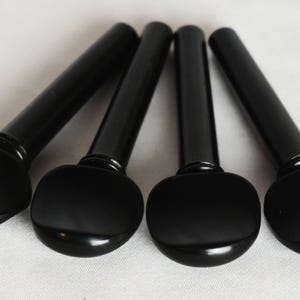 Puede incluir: Cuatro clavijas de afinación de violín negras dispuestas en diagonal sobre una superficie blanca. Cada clavija tiene una cabeza redondeada y ovalada y un eje cilíndrico. Las clavijas son de material liso y brillante.