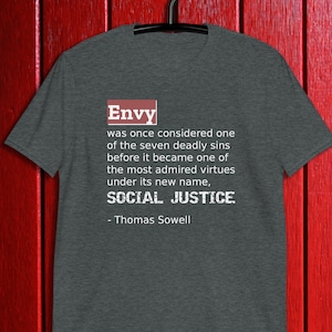 May include: A dark grey t-shirt featuring the word "Envy" in a red box, and a quote about social justice by Thomas Sowell in white text. The t-shirt is displayed on a black hanger against a red wooden background.