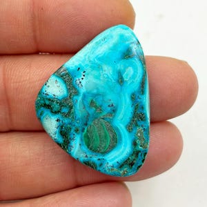 May include: A polished gemstone with a triangular shape. The stone features a vibrant mix of turquoise and teal hues, with darker green and black veining patterns. The surface is smooth and reflective, showcasing the natural variations in color and texture.