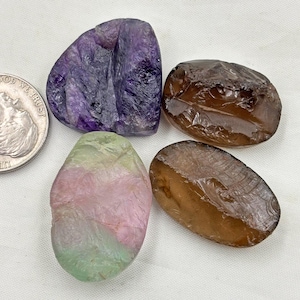 Pode incluir: Quatro pedras preciosas polidas em tons de roxo, castanho, rosa e verde. As pedras preciosas têm formato irregular e textura áspera. Uma moeda de prata está ao lado das pedras preciosas para comparação de tamanho.