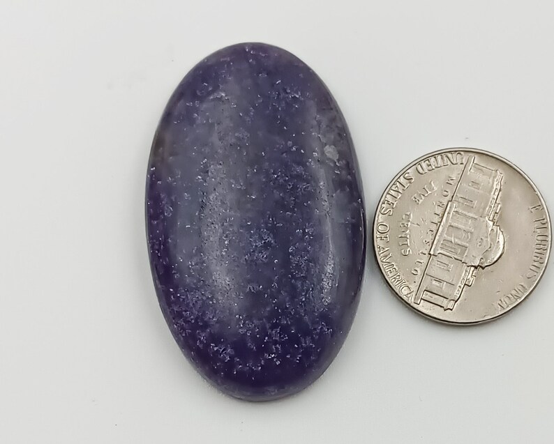 K&ouml;nnte beinhalten: Ein ovaler Cabochon-Edelstein in Lila und Grau mit glitzernder Oberfl&auml;che. Der Edelstein ist etwa so gro&szlig; wie eine deutsche Zehn-Cent-M&uuml;nze.