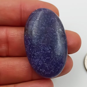K&ouml;nnte beinhalten: Ein glatter, ovaler, violetter Edelstein mit einer funkelnden, glitzernden Textur. Der Edelstein ist etwa so gro&szlig; wie eine deutsche 2-Euro-M&uuml;nze.