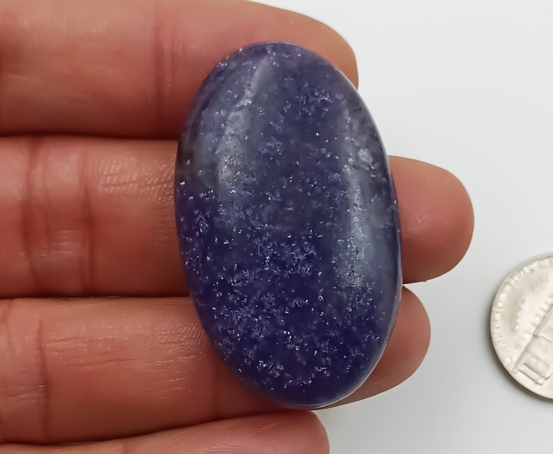 K&ouml;nnte beinhalten: Ein glatter, ovaler, violetter Edelstein mit einer funkelnden, glitzernden Textur. Der Edelstein ist etwa so gro&szlig; wie eine deutsche 2-Euro-M&uuml;nze.