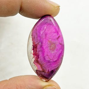 Pu&ograve; includere: Una gemma lucida di forma ovale, con un vivace centro magenta e un bordo traslucido bianco sporco. La pietra mostra venature naturali e variazioni di colore, tenuta tra le dita.