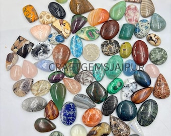 May include: A collection of polished gemstones in various shapes and colors, including green, orange, blue, and brown. The gemstones are scattered across a white surface, with the text "CRAFT GEMS JAIPUR" visible.