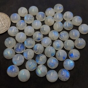 Op de afbeelding: Een verzameling van 35 ronde, witte maansteen cabochons met iriserende blauwe en groene schittering. De cabochons zijn ongeveer 10 mm in diameter.