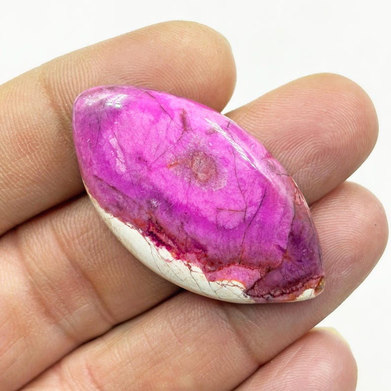 Pu&ograve; includere: Una gemma lucida di forma ovale, con una vibrante tonalit&agrave; magenta, caratterizzata da venature intricate e una base bianca crema. La superficie della pietra &egrave; liscia e riflettente, mostrando un motivo naturale e organico. La gemma &egrave; tenuta in una mano.