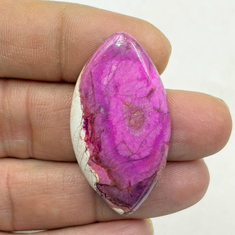 Pu&ograve; includere: Una gemma lucida di forma ovale, con un vibrante colore magenta, venata di viola pi&ugrave; scuro e un bordo bianco crema. La pietra &egrave; tenuta in mano, mostrando i suoi motivi naturali.