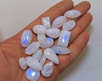 Op de afbeelding: Een verzameling witte maansteen cabochons met iriserende blauwe flitsen. De stenen zijn in verschillende vormen en maten, waaronder ovaal, peervormig en rechthoekig.