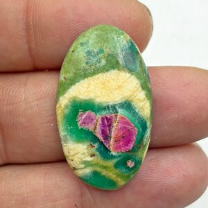 Könnte beinhalten: Ovaler Edelstein mit einer lebendigen Farbpalette. Der Stein hat eine Basis aus Grün- und Gelbtönen mit einem auffälligen Zentrum aus tiefrosa und roten kristallinen Formationen. Der Edelstein wird in einer Hand gehalten.