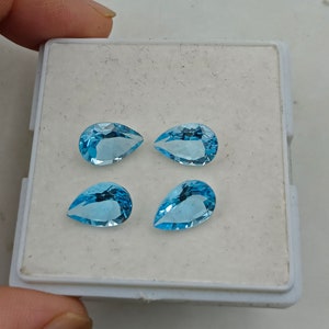 May include: Four pear-shaped blue topaz gemstones in a white display box. The gemstones are faceted and have a light blue color.