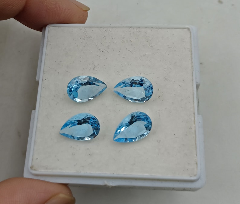 May include: Four pear-shaped blue topaz gemstones in a white display box. The gemstones are faceted and have a light blue color.
