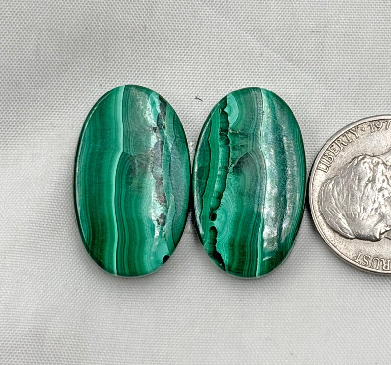 May include: Two oval-shaped malachite stones with a vibrant green color and unique swirling patterns. The stones are displayed next to a US quarter for size comparison. The malachite has dark green and black veining.