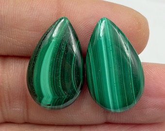 May include: Two teardrop-shaped malachite gemstones. The gemstones are a vibrant green with darker green swirling patterns. The polished stones are held in a hand, showcasing their natural beauty and unique veining.