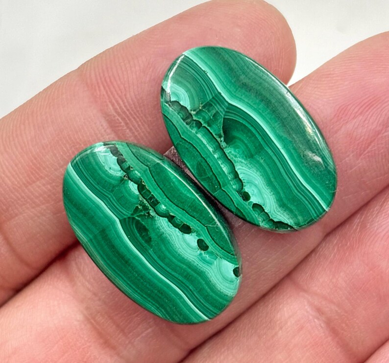 May include: Two polished oval malachite gemstones. The gemstones are a vibrant green with darker green, swirling patterns. The stones are held in a hand, showcasing their natural beauty and unique veining.