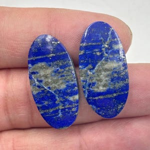 Könnte beinhalten: Zwei ovale Lapislazuli-Cabochons mit einem blauen und silbernen Farbmuster. Die Steine haben eine glatte, polierte Oberfläche.