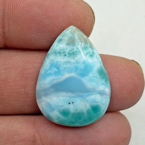 May include: A teardrop-shaped gemstone with a light blue and white pattern, reminiscent of ocean waves. The stone has a polished surface and a mix of turquoise and white hues. The gemstone is held between two fingers.
