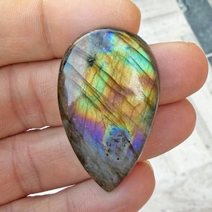 May include: A teardrop-shaped labradorite gemstone with a polished surface. The stone displays iridescent colors including blue, green, yellow, and purple. The gemstone is held in a hand, showcasing its natural beauty and unique color play.