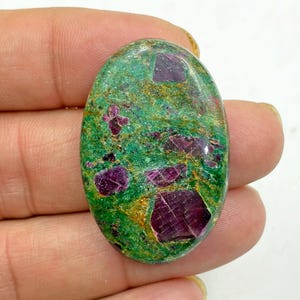 May include: Oval-shaped gemstone with a green base color and vibrant ruby inclusions. The ruby crystals are deep red and scattered throughout the stone, creating a striking contrast. The gemstone is polished and smooth.