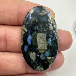 Könnte beinhalten: Ein ovaler Cabochon-Edelstein in Schwarz, Grün und Blau mit einem gesprenkelten Muster. Der Edelstein ist etwa 5 cm lang und 2,5 cm breit.
