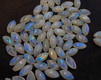 Puede incluir: Una colección de cabochones de piedra lunar de forma ovalada, de color blanco lechoso y con destellos iridiscentes azules y verdes. Las piedras están dispuestas en un montón suelto sobre un fondo oscuro.