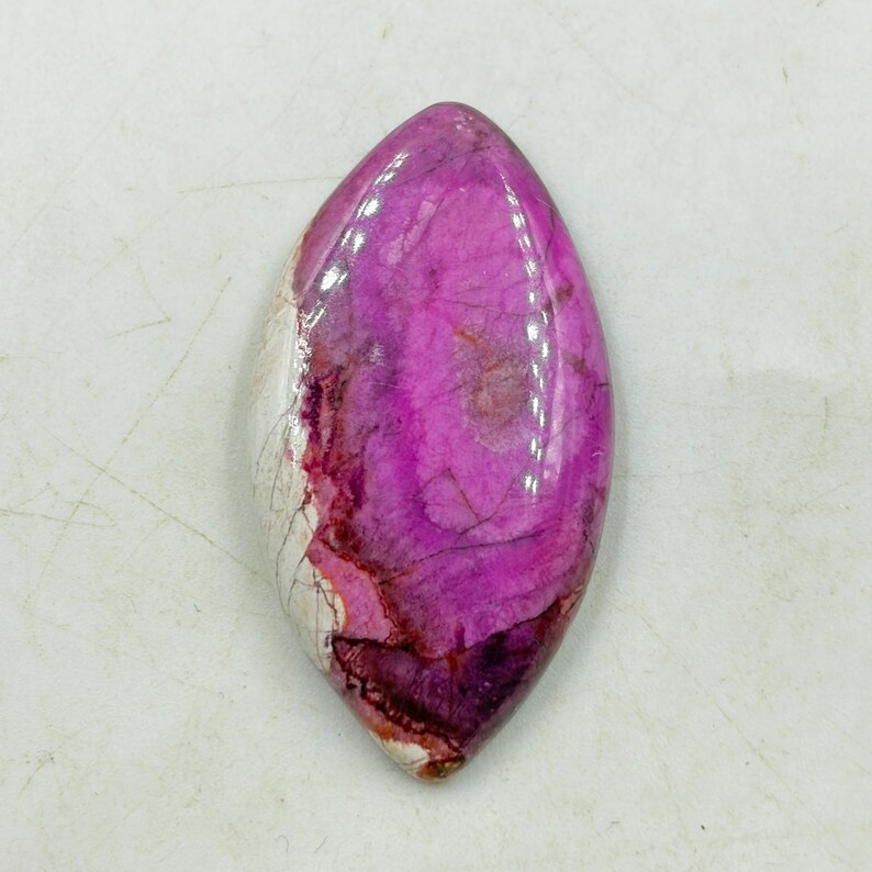 Pu&ograve; includere: Una gemma lucida e allungata con una vibrante tonalit&agrave; magenta, venata di tonalit&agrave; pi&ugrave; scure di rosso e viola. La pietra ha una superficie liscia e lucida e una sezione di marmorizzazione bianca e crema. La forma generale ricorda una lacrima o un occhio.