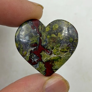 Pode incluir: Uma pedra em forma de coração com uma superfície polida. A pedra apresenta uma mistura de cores, incluindo verde, vermelho e preto. O coração é segurado entre dois dedos.