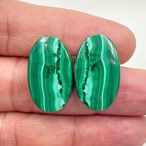 May include: Two oval-shaped malachite gemstones. The polished stones display a vibrant green color with intricate swirling patterns. The gemstones are held between fingers, showcasing their size and detail. The malachite has a natural, organic appearance.