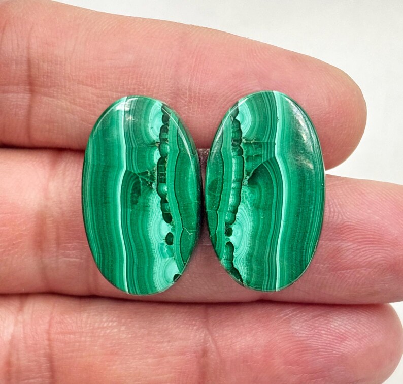 May include: Two oval-shaped malachite gemstones. The polished stones display a vibrant green color with intricate swirling patterns. The gemstones are held between fingers, showcasing their size and detail. The malachite has a natural, organic appearance.